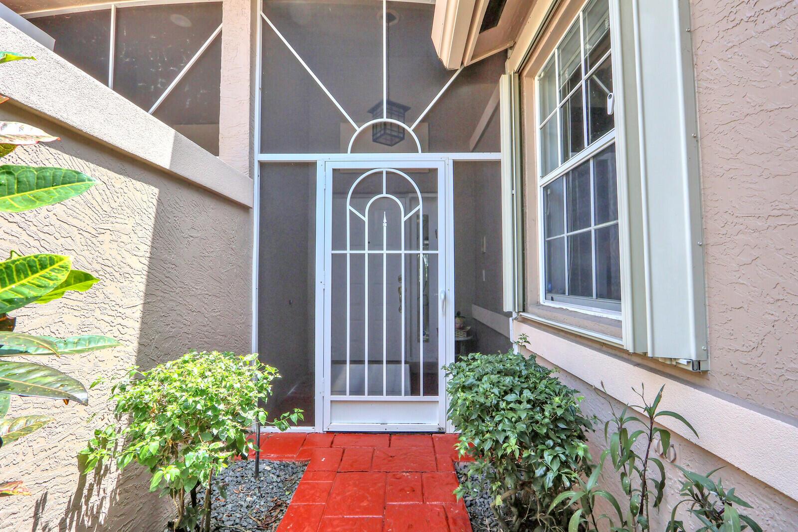 A white screen door leads to an entryway with a red tiled path and landscaping — 9531 Crescent View Drive N, Boynton Beach FL 33437, BOYNTON BEACH, FL