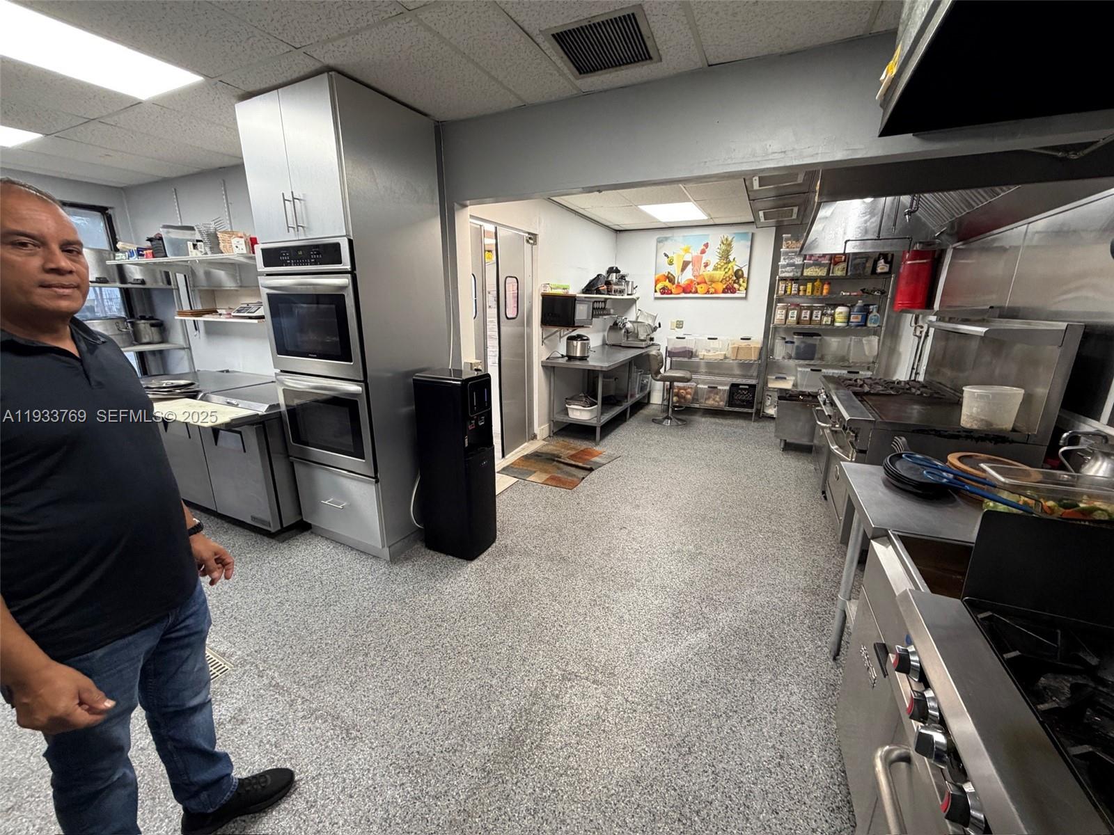 A man stands in a commercial kitchen featuring stainless steel appliances and grey flooring — for sale SW restaurant, Miami FL 33166, MIAMI, FL