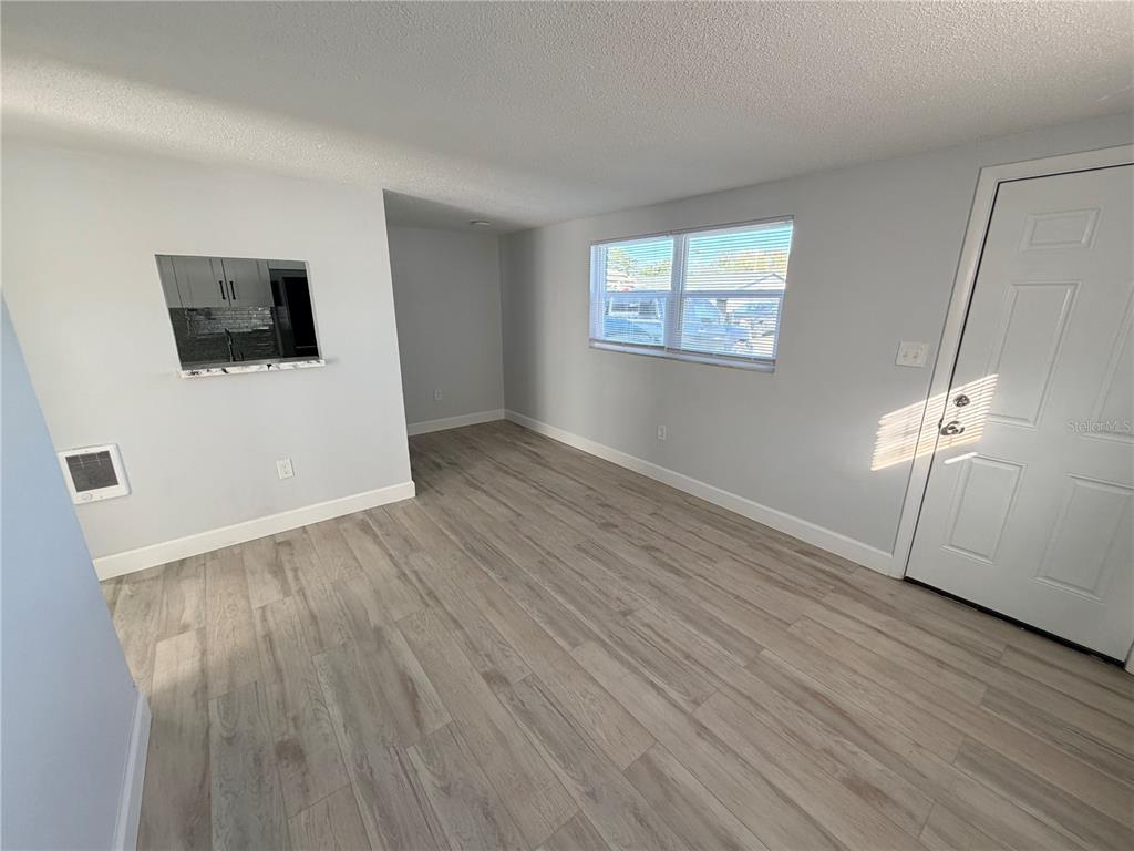 An empty room features light wood-look flooring, gray walls, a window, and a kitchen pass-through — 1101 E 140TH AVENUE, TAMPA, FL