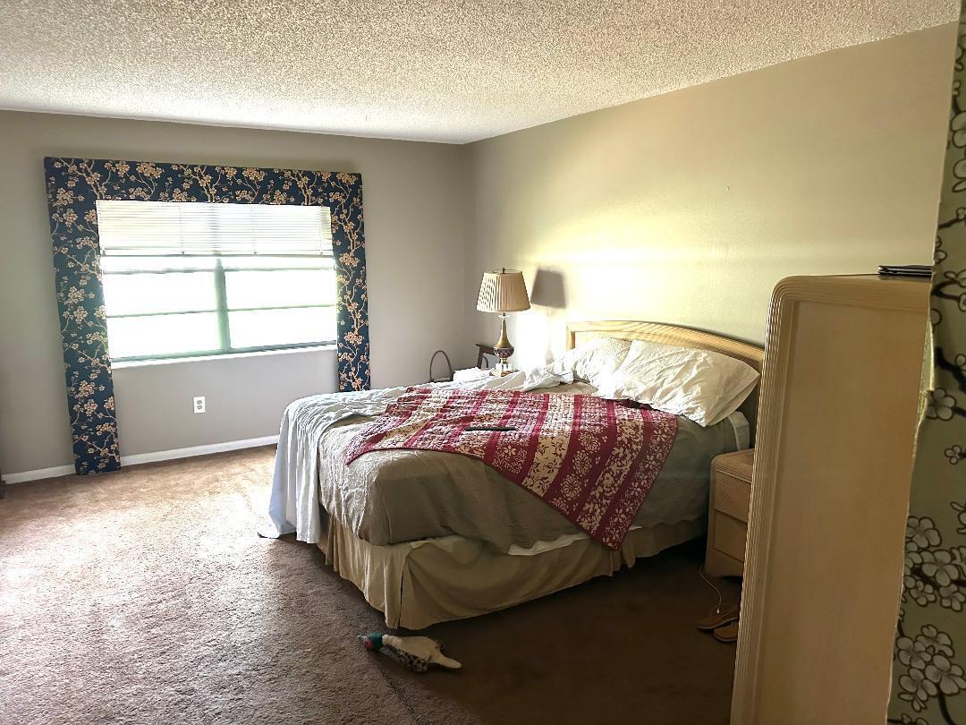 A bedroom features a bed with a patterned quilt, a window with floral curtains, and carpeting — 21848 Arriba Real # A, Boca Raton FL 33433, BOCA RATON, FL