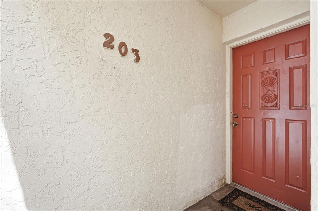 A red front door with a decorative panel is adjacent to a wall labeled 203 — 7500 40TH AVENUE W UNIT 203, BRADENTON, FL