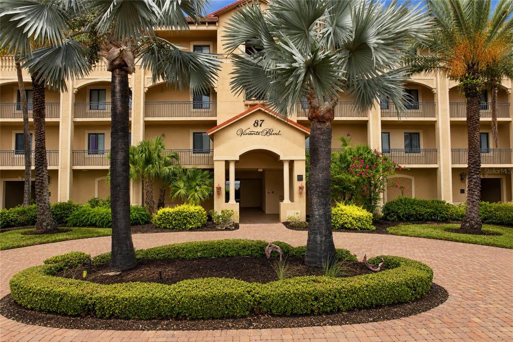 A multi-story tan apartment building features a central entrance with palm trees and landscaping — 87 VIVANTE BOULEVARD UNIT 206, PUNTA GORDA, FL