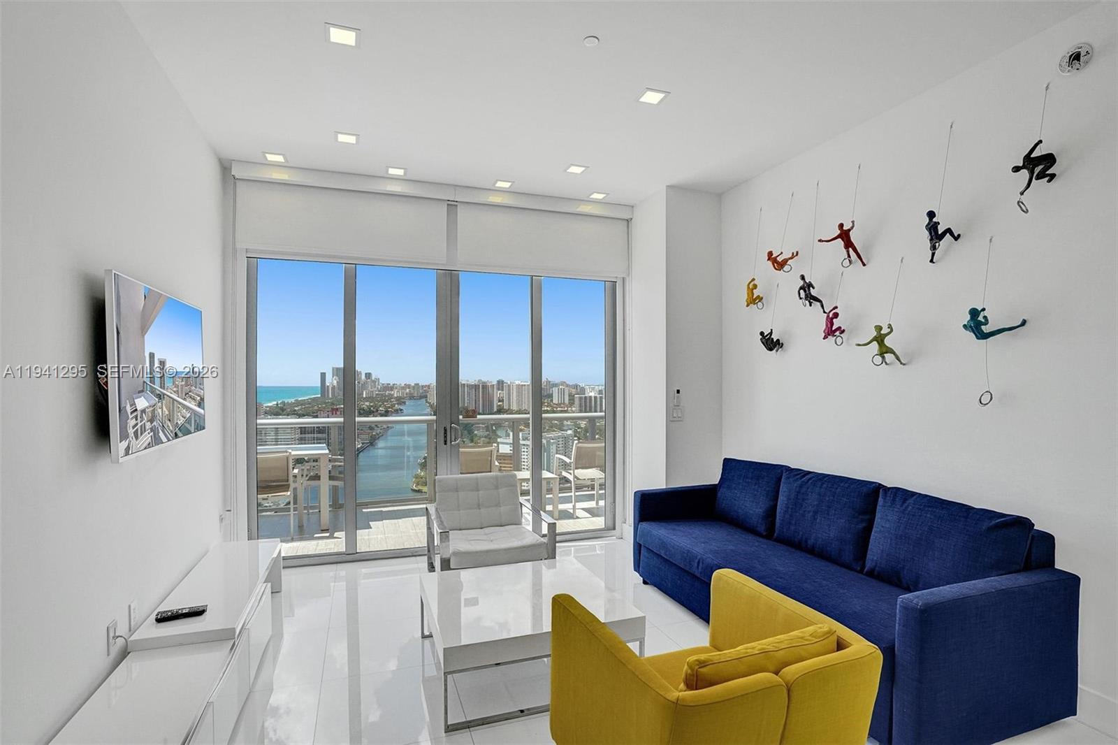A modern living room features a blue sofa, yellow armchair, and wall-mounted climbing figure sculptures — 2600 E Hallandale Beach Blvd # T3303, Hallandale Beach FL 33009, HALLANDALE BEACH, FL