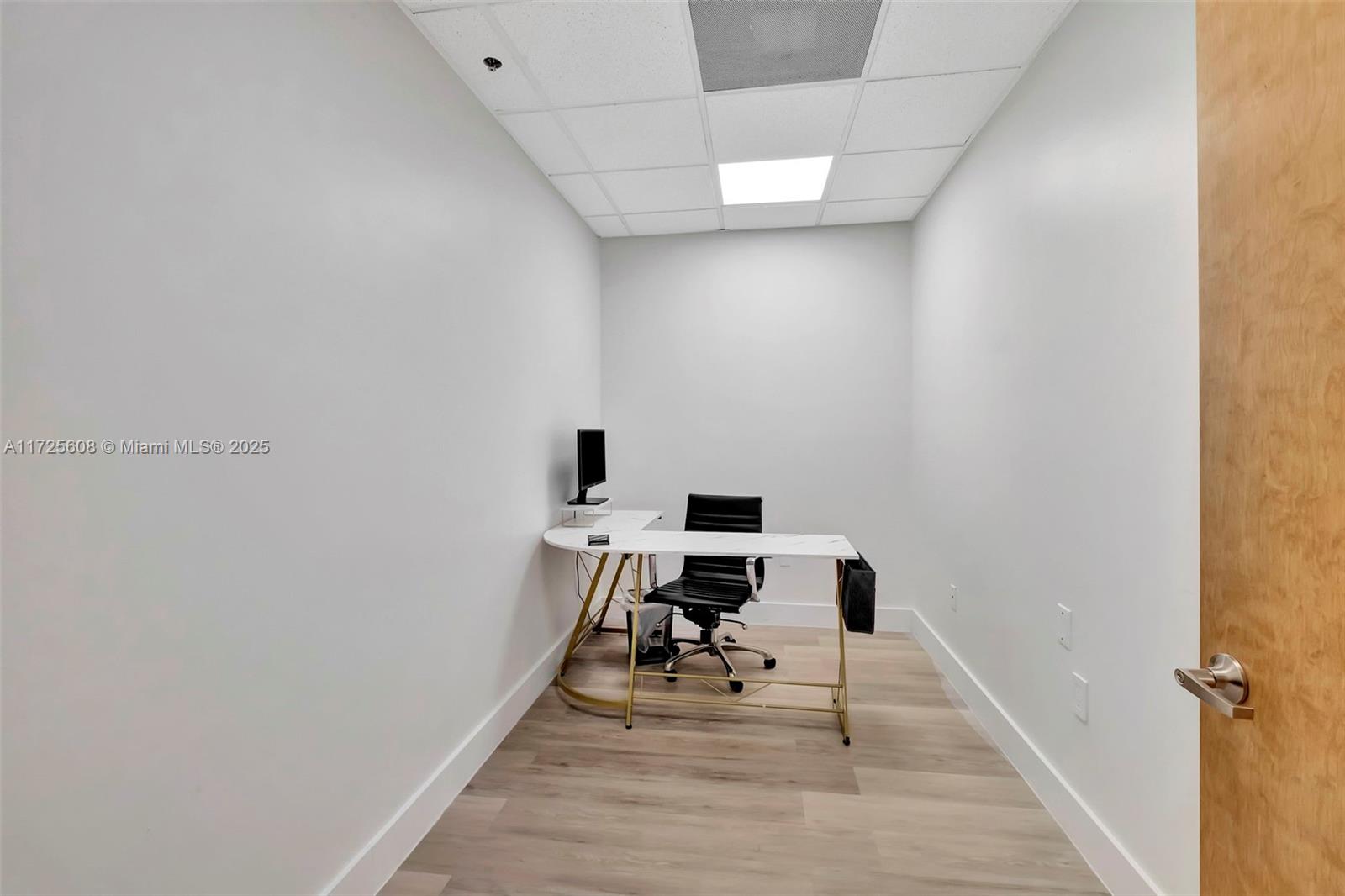 A small, minimalist office features a white L-shaped desk, a black chair, and light wood flooring — 12550 Biscayne Blvd # 15, North Miami FL 33181, NORTH MIAMI, FL