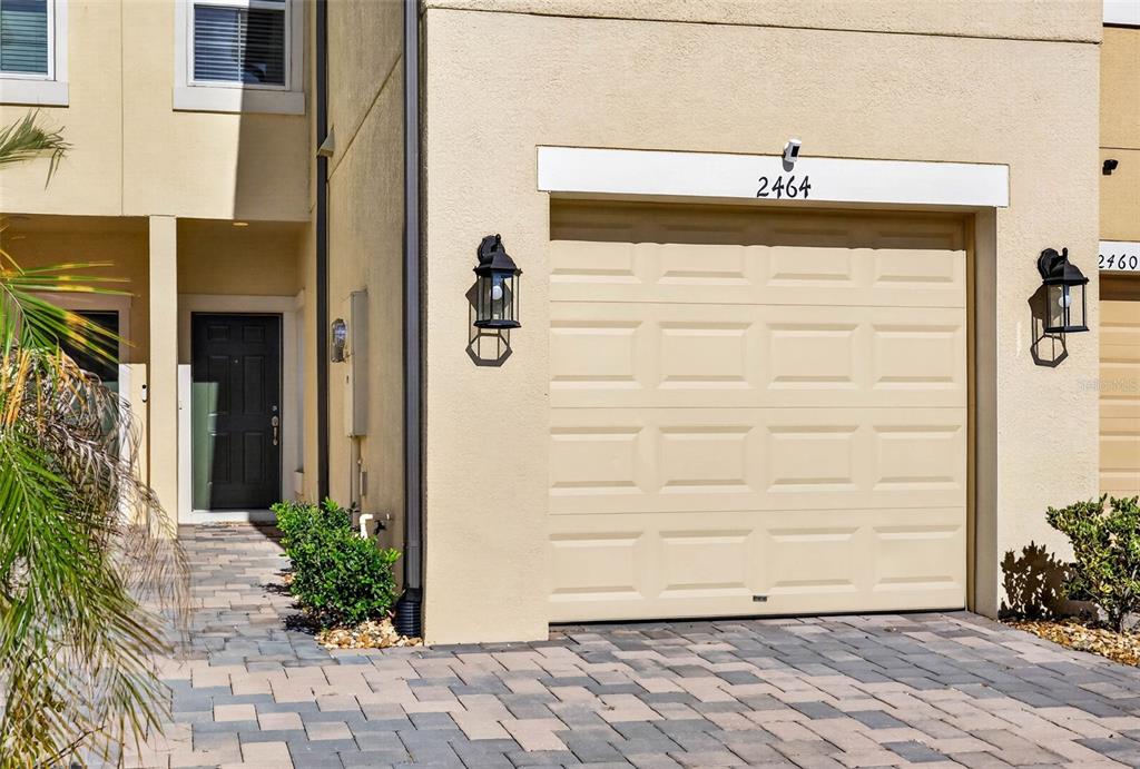 A beige townhouse exterior features a single-car garage, a dark front door, and paver driveway — 2464 STAPLEFORD PLACE, WESLEY CHAPEL, FL