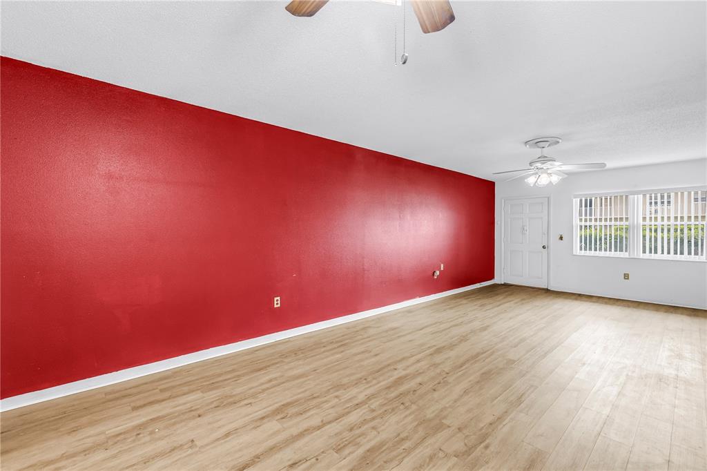 An empty living room features a prominent red accent wall, light wood-look flooring, and windows — 5729 13TH AVENUE N UNIT 102D, ST PETERSBURG, FL