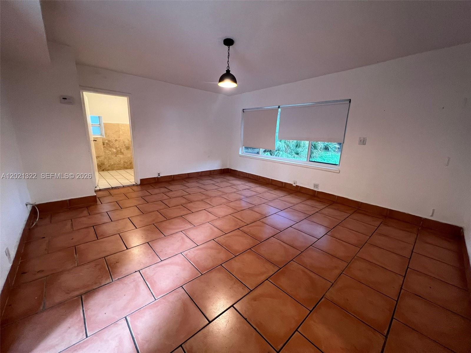 An empty room features terracotta tile flooring, white walls, a pendant light, and a window — 644 NE 82nd Ter # 1, Miami FL 33138, MIAMI, FL