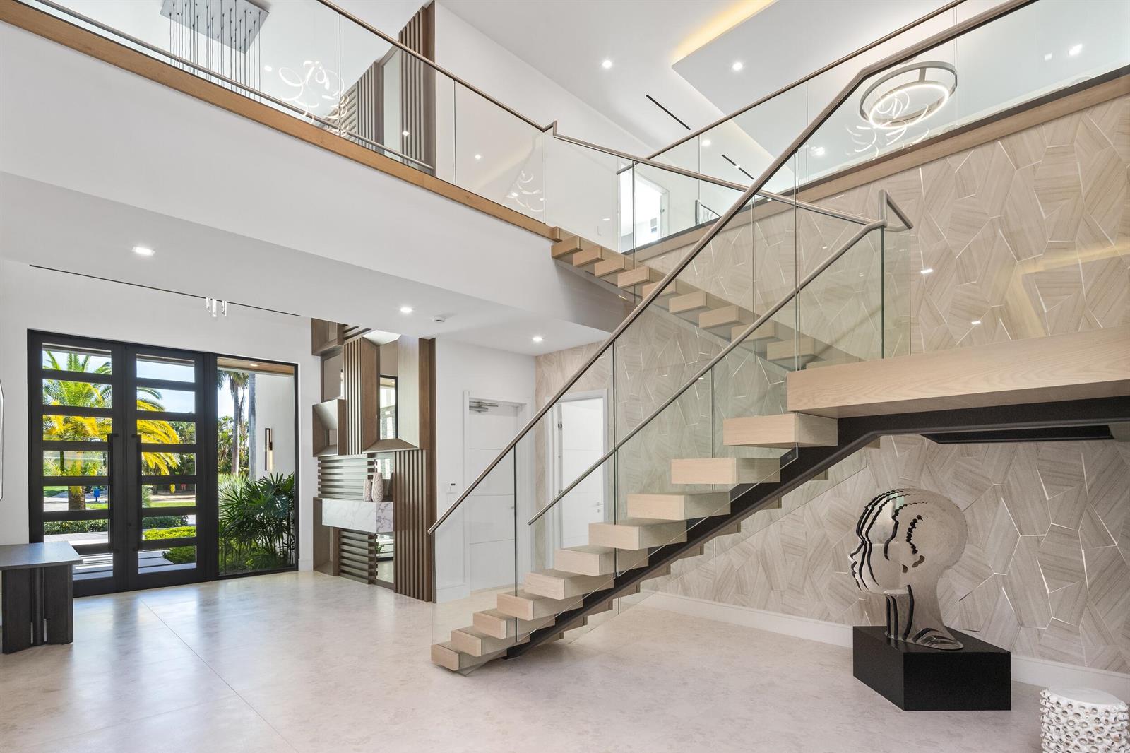 A modern foyer features a floating staircase with glass railings and a geometric tiled wall — 217 Thatch Palm Drive, Boca Raton FL 33432, BOCA RATON, FL
