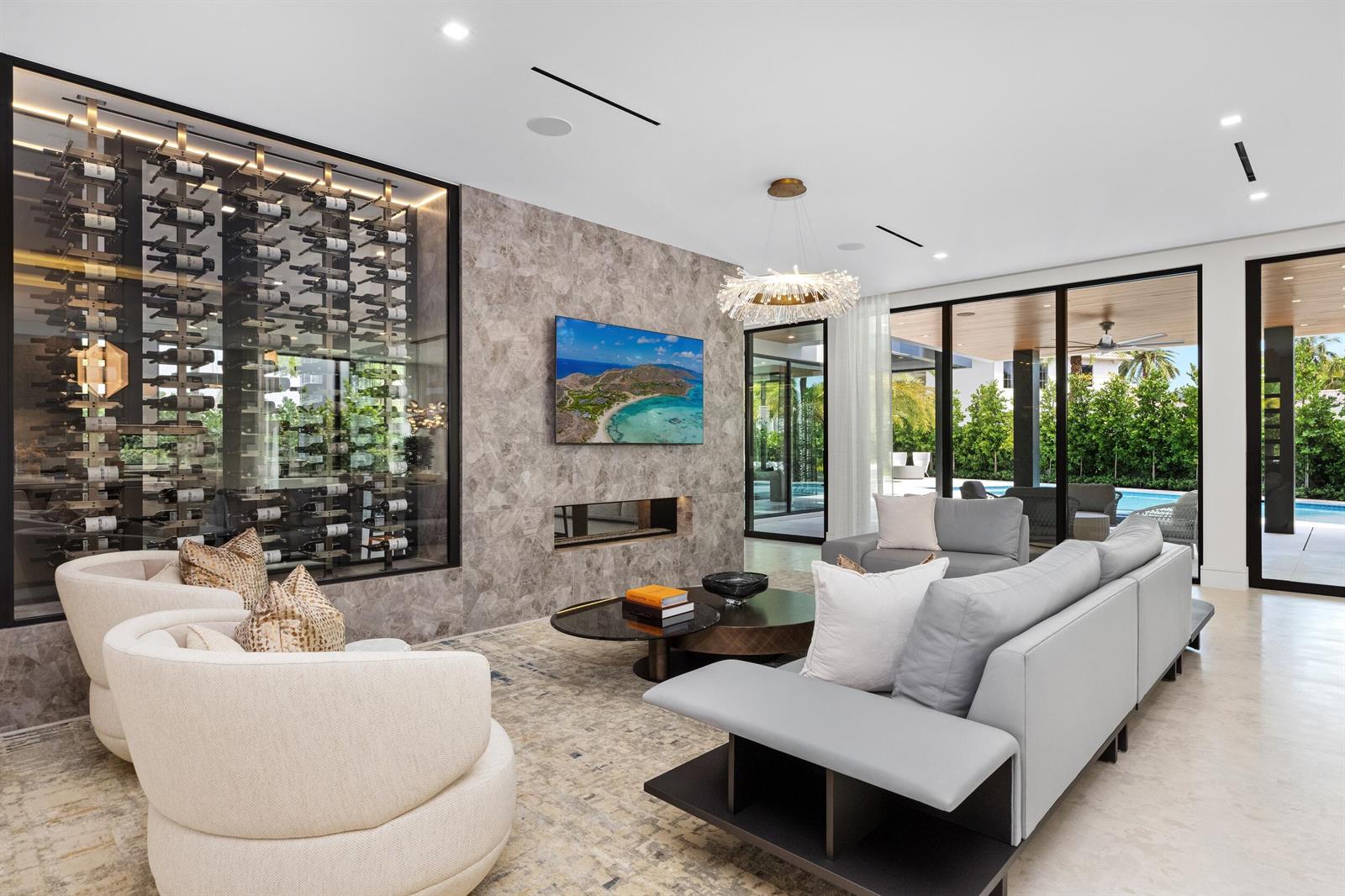 A modern living room features a glass wine wall, stone fireplace, and floor-to-ceiling windows — 217 Thatch Palm Drive, Boca Raton FL 33432, BOCA RATON, FL