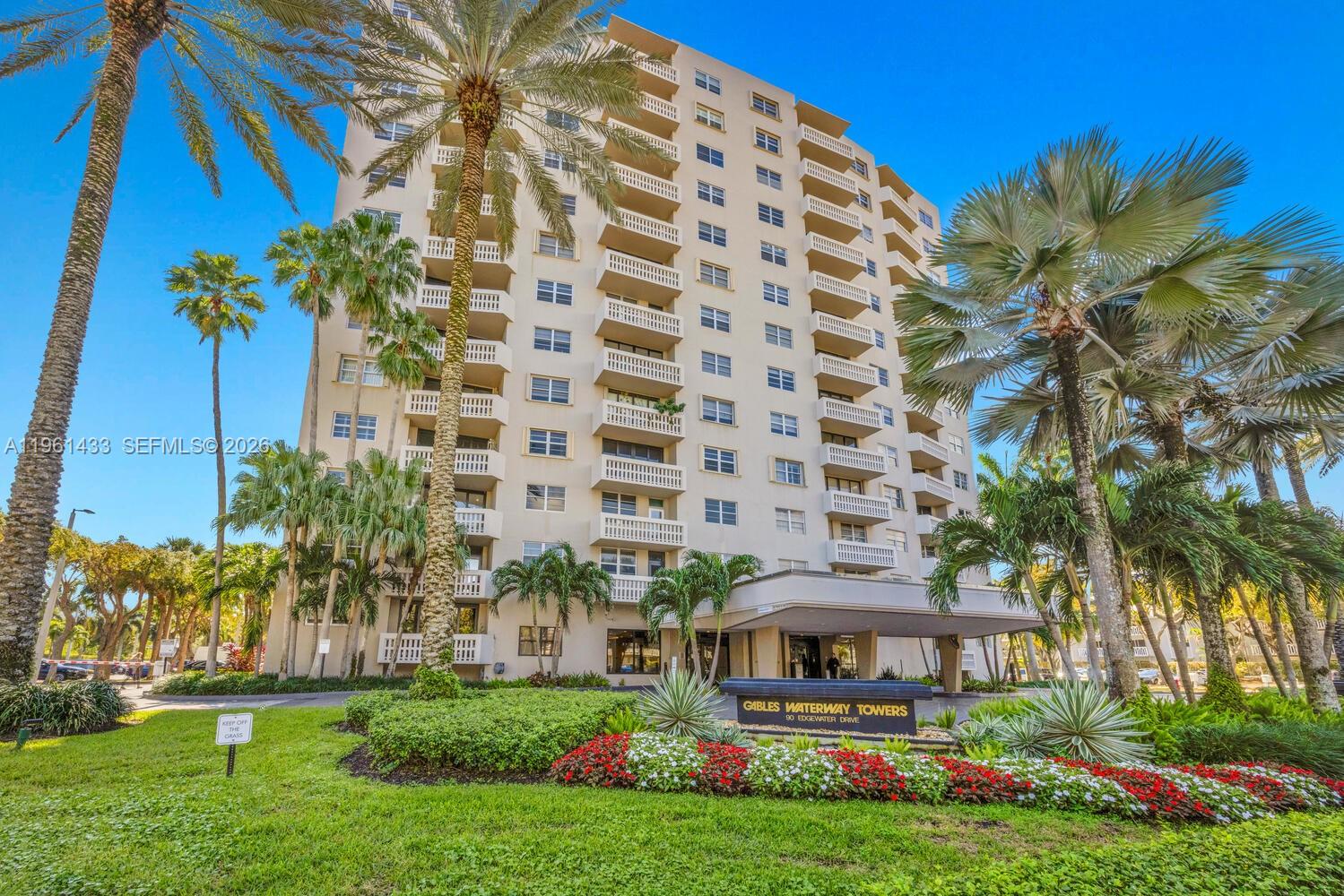 A tall residential building with balconies stands behind a landscaped garden and palm trees — 90 Edgewater Dr, Coral Gables FL 33133, CORAL GABLES, FL