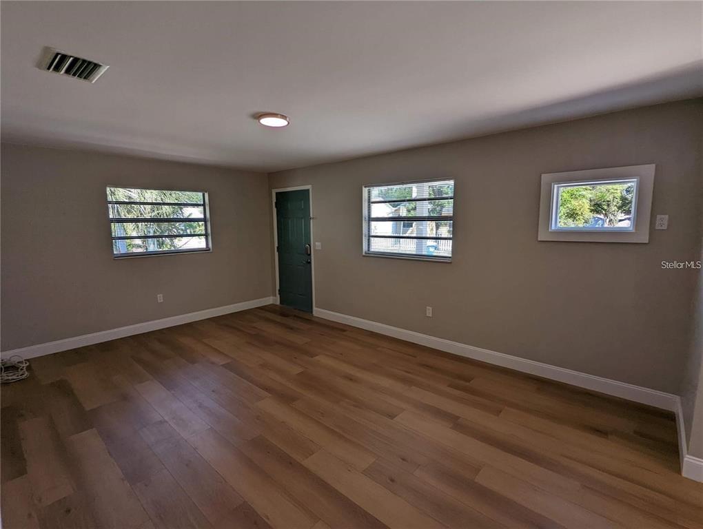 An empty room features light wood-look flooring, gray walls, three windows, and a dark door — 1920/1922 SPRINGTIME AVENUE, CLEARWATER, FL