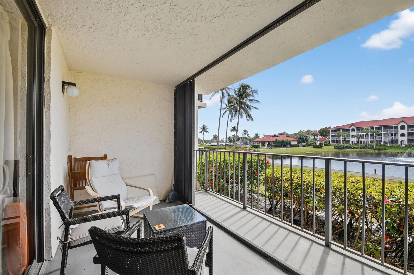 A covered balcony features patio furniture overlooking a lake and multi-story residential buildings — 7030 Half Moon Circle # 117, Hypoluxo FL 33462, HYPOLUXO, FL