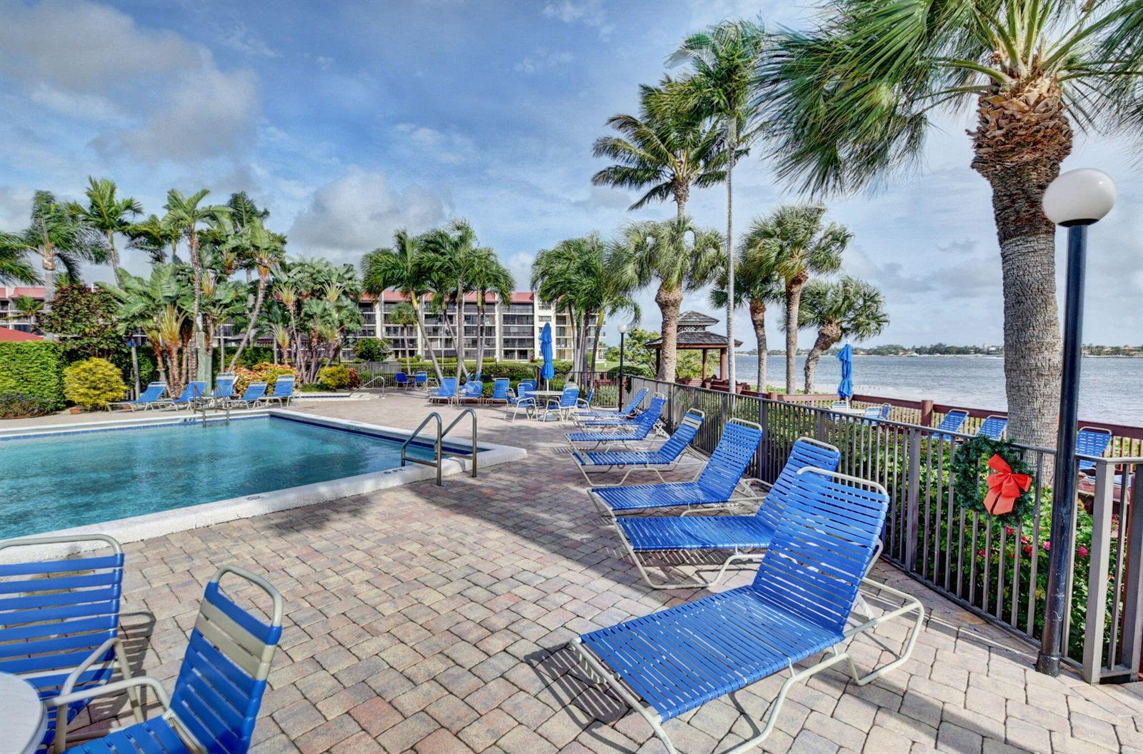 A swimming pool area with blue lounge chairs, palm trees, and a view of the water — 7030 Half Moon Circle # 117, Hypoluxo FL 33462, HYPOLUXO, FL