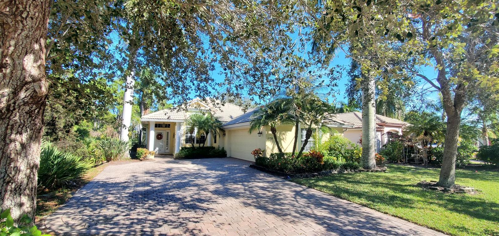 A yellow single-story house with a paved driveway, palm trees, and a tiled roof — 1128 NW Mossy Oak Way, Jensen Beach FL 34957, JENSEN BEACH, FL