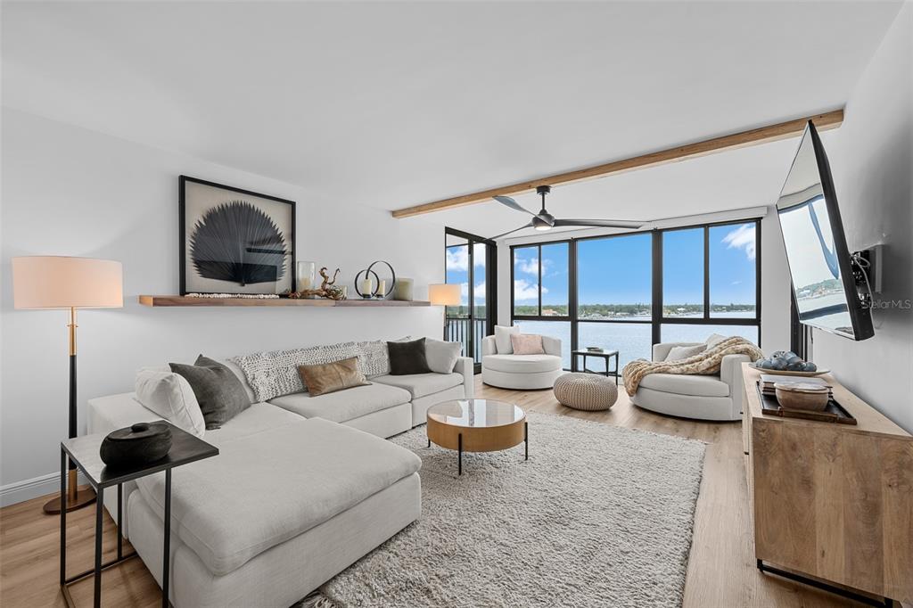 A modern living room features a white sectional sofa, wood-toned furniture, and floor-to-ceiling waterfront windows — 7100 SUNSHINE SKYWAY LANE S UNIT 701, ST PETERSBURG, FL