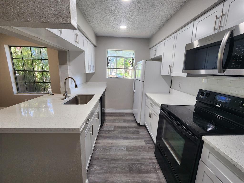 A galley kitchen features white cabinets, quartz countertops, stainless steel appliances, and gray wood-look flooring — 200 AFTON SQUARE UNIT 108, ALTAMONTE SPRINGS, FL