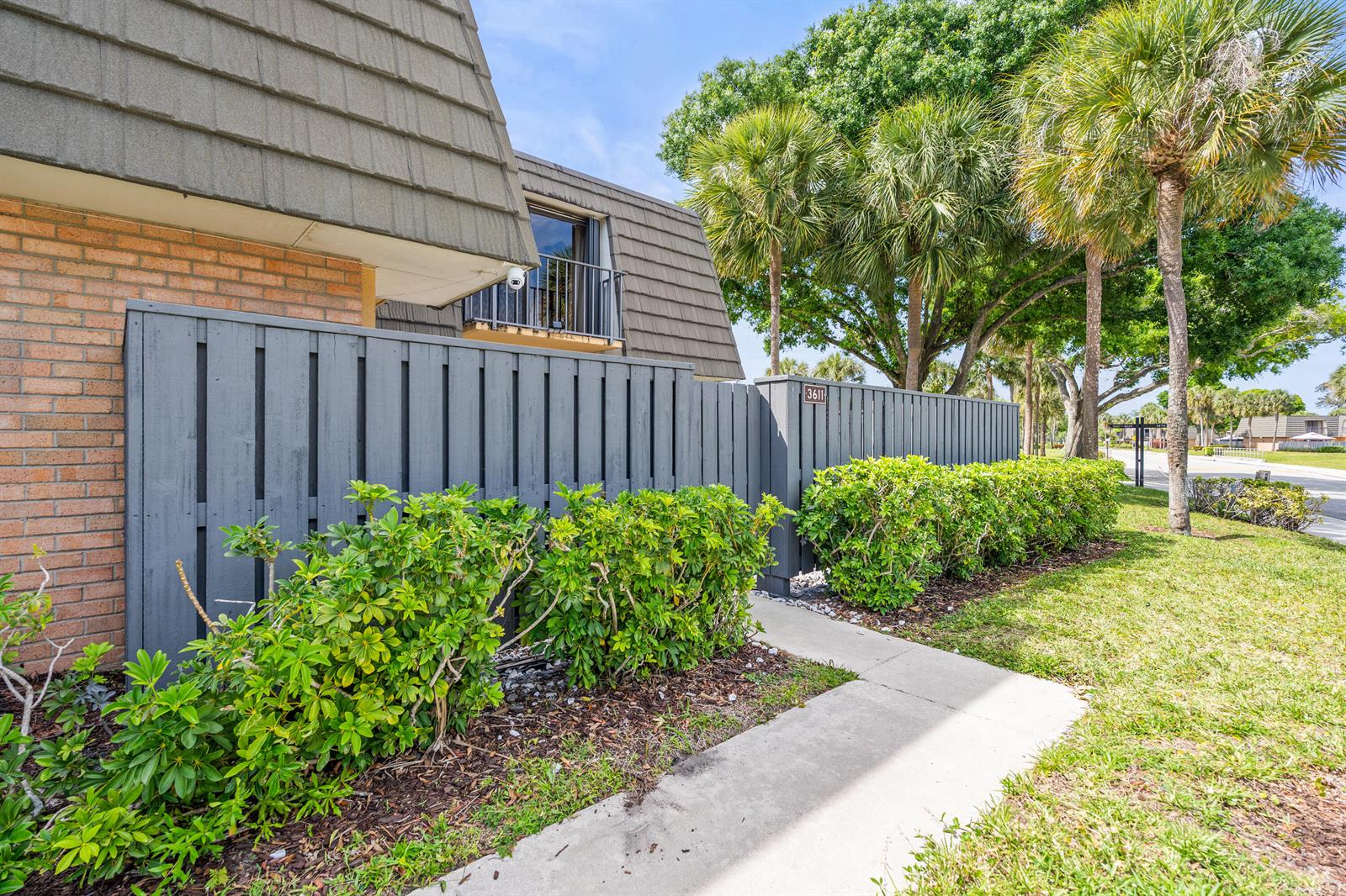 A gray wooden privacy fence lines a concrete walkway beside a brick building with palm trees — 3611 36th Way, West Palm Beach FL 33407, WEST PALM BEACH, FL