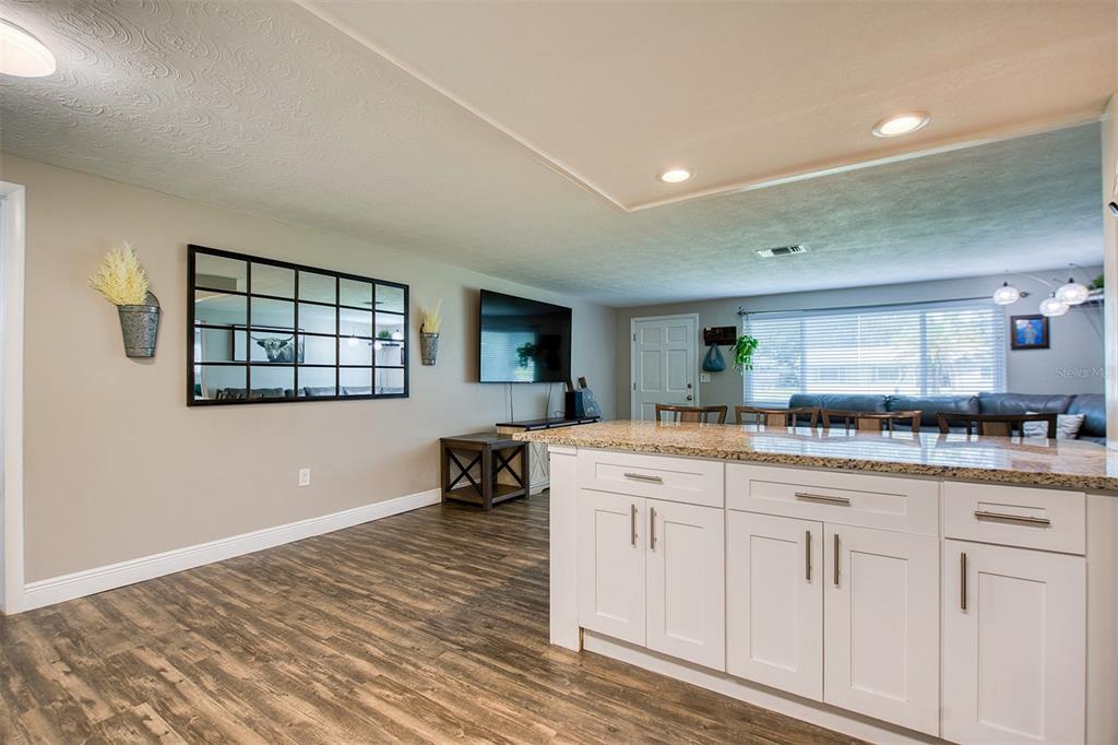 A kitchen island with white cabinets and granite countertops overlooks a living area with wood-look flooring — 1440 WISCONSIN AVENUE, PALM HARBOR, FL
