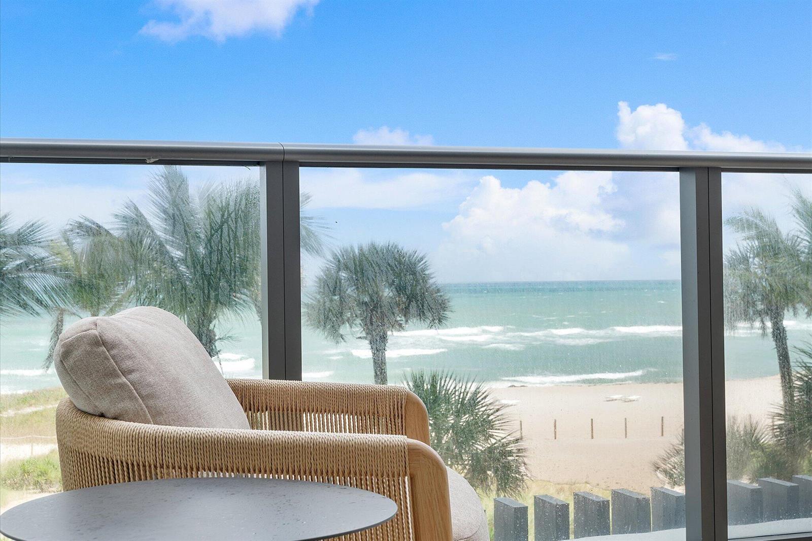 A balcony with a woven chair and glass railing overlooks a beach and ocean — 1116 N Ocean Boulevard # 304, Pompano Beach FL 33062, POMPANO BEACH, FL