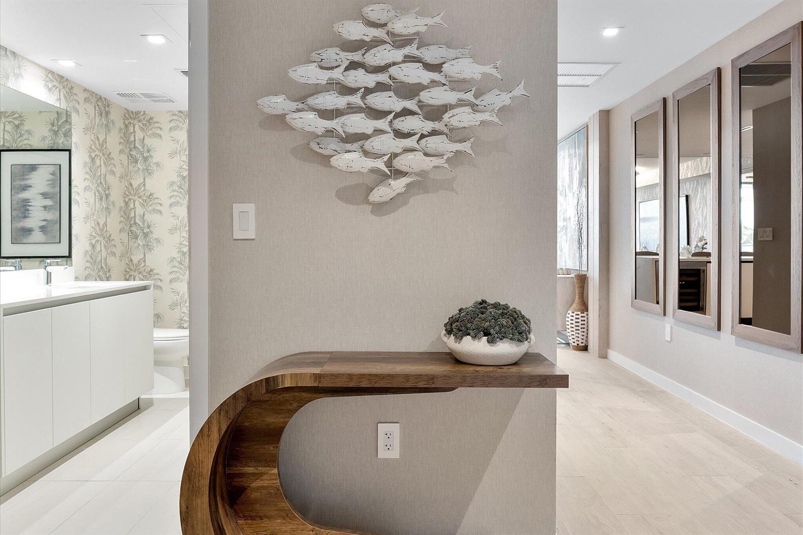 A modern hallway features a curved wooden console table, a fish wall sculpture, and mirrors — 1116 N Ocean Boulevard # 304, Pompano Beach FL 33062, POMPANO BEACH, FL