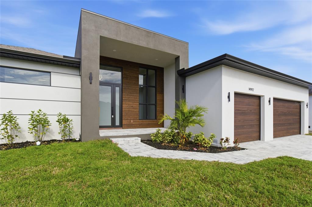 A modern single-story house exterior features a gray entryway, wood-look garage doors, and white walls — 14192 FORT MYERS AVENUE, PORT CHARLOTTE, FL