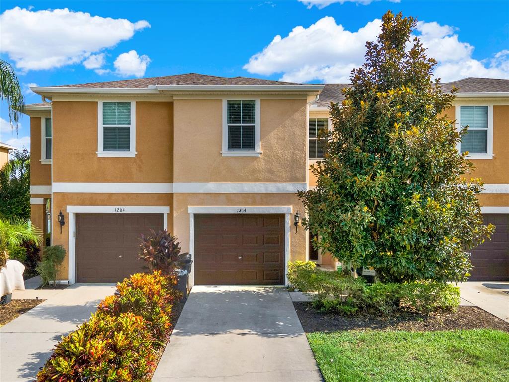 A two-story townhouse exterior features tan stucco walls, brown garage doors, and a large tree — 1214 CHELSEA DRIVE, DAVENPORT, FL