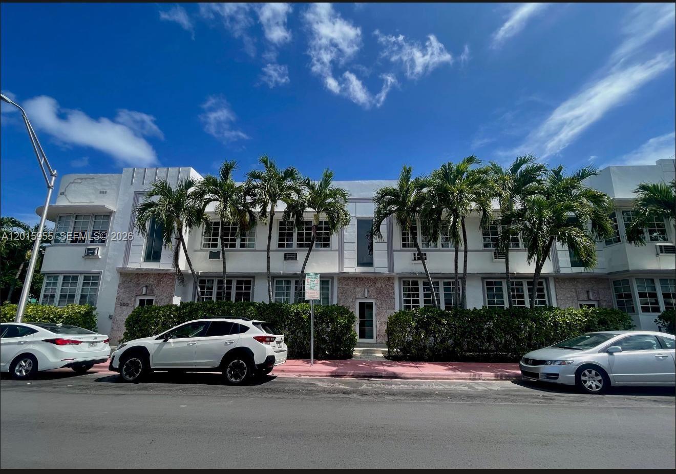 A two-story white Art Deco apartment building with palm trees and parked cars in front — MIAMI BEACH, FL