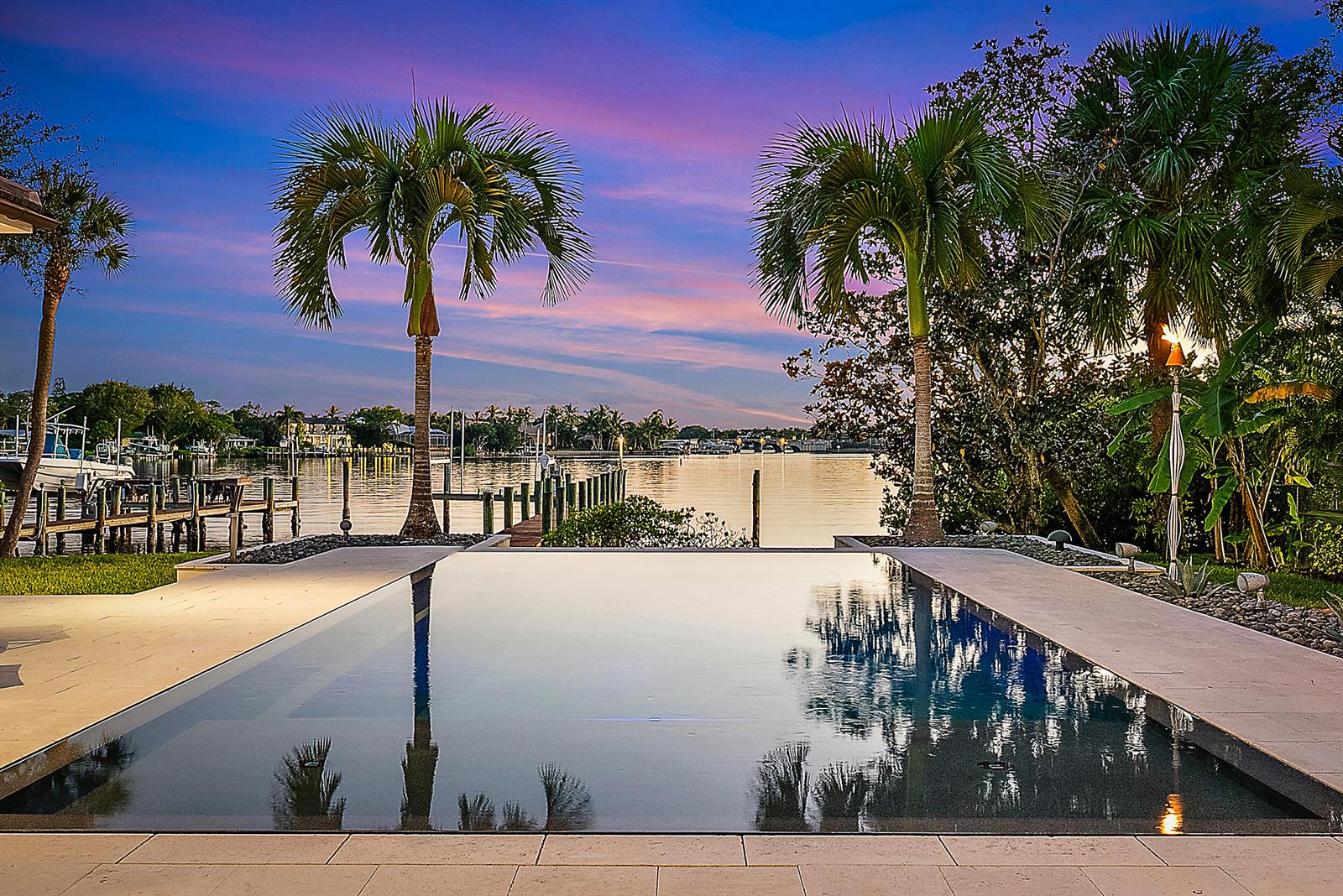 An infinity pool overlooks a calm waterway with boat docks and palm trees under blue skies — 19651 Riverside Drive, Jupiter FL 33469, JUPITER, FL