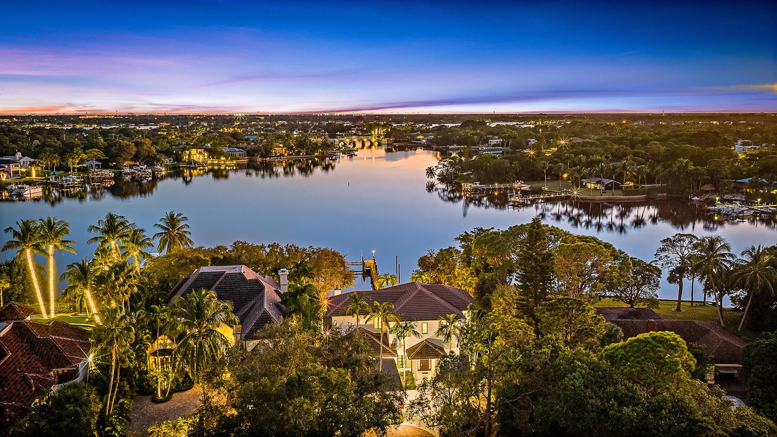 An aerial view of a waterfront estate at dusk with lush landscaping and a dock — 19651 Riverside Drive, Jupiter FL 33469, JUPITER, FL