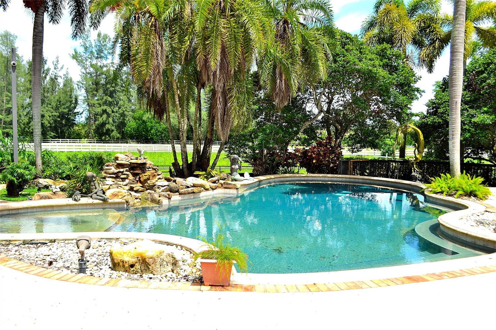 A backyard swimming pool features a rock waterfall, palm trees, and a surrounding patio area — 6200 SW 183rd Way, Southwest Ranches FL 33331, SOUTHWEST RANCHES, FL