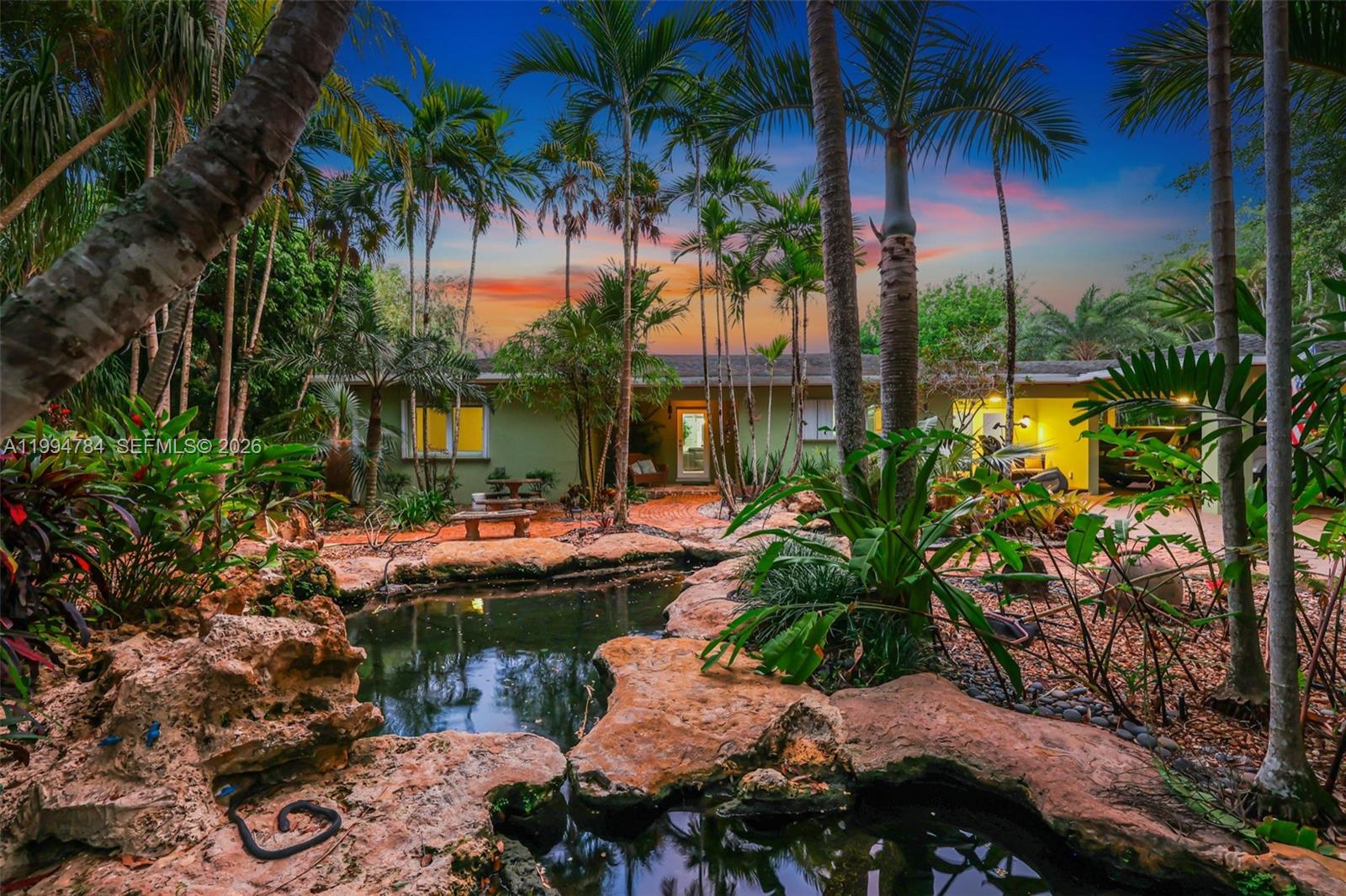 A backyard features a rock-lined pond, lush tropical landscaping, and a single-story house at dusk — 9234 SW 112th St, Miami FL 33176, MIAMI, FL
