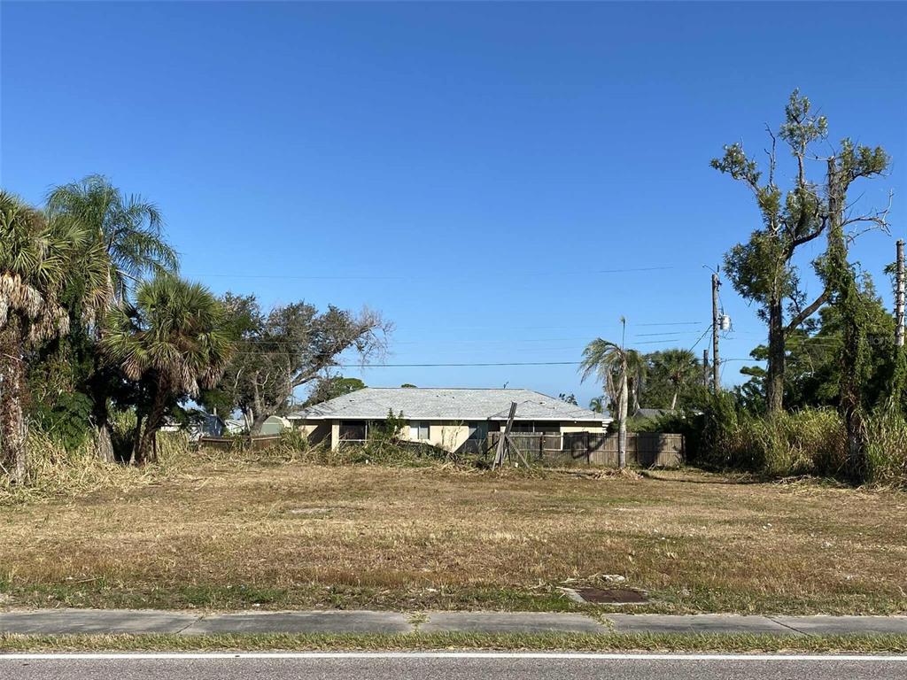 A vacant, grassy lot sits in front of a single-story house with a gray roof — 7439 SAN CASA DRIVE, ENGLEWOOD, FL