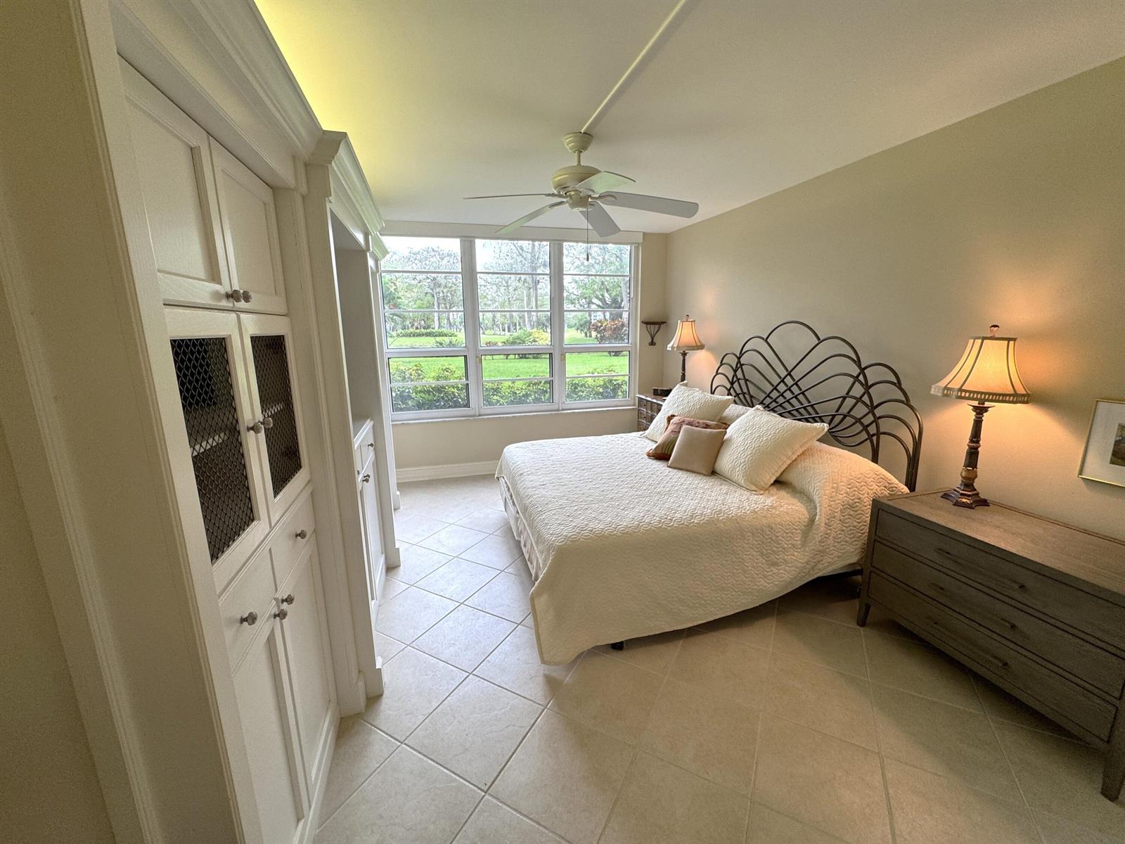 A bedroom features a metal headboard, white cabinetry, tile flooring, and a large window view — 625 Oaks Drive # 106, Pompano Beach FL 33069, POMPANO BEACH, FL