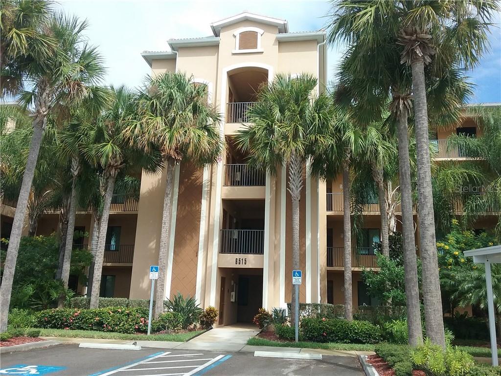 A multi-story tan apartment building features balconies, palm trees, and a handicap-accessible parking space — 6515 GRAND ESTUARY TRAIL UNIT 402, BRADENTON, FL