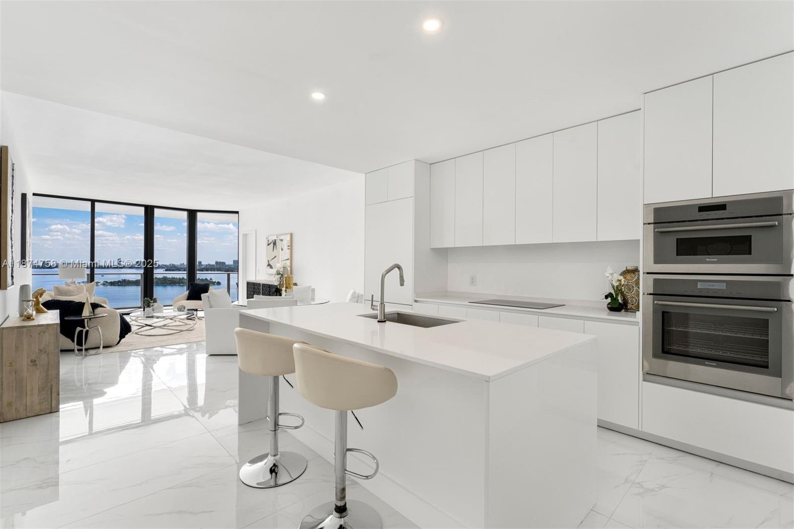A modern white kitchen with an island and bar stools overlooks a waterfront living area — 700 NE 24th St # 1005, Miami FL 33137, MIAMI, FL