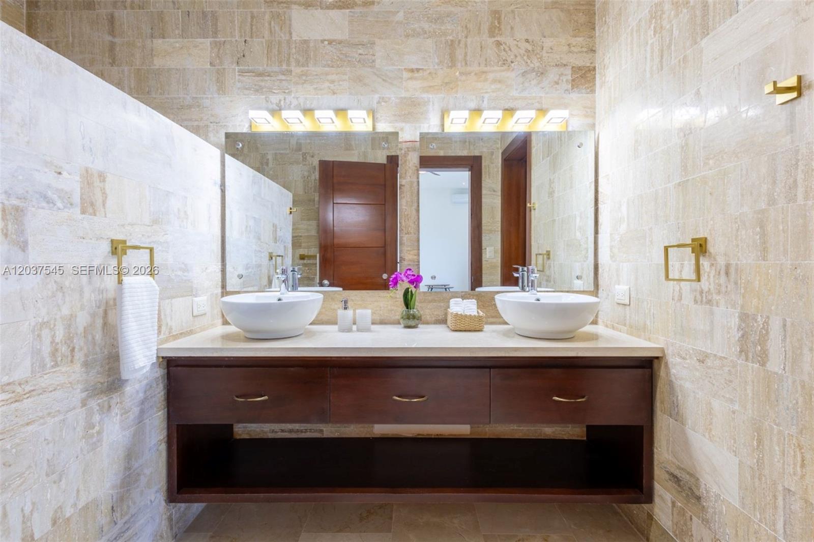 A bathroom features a double vanity with vessel sinks, gold hardware, and beige stone tiling — 0 la palmass, FL