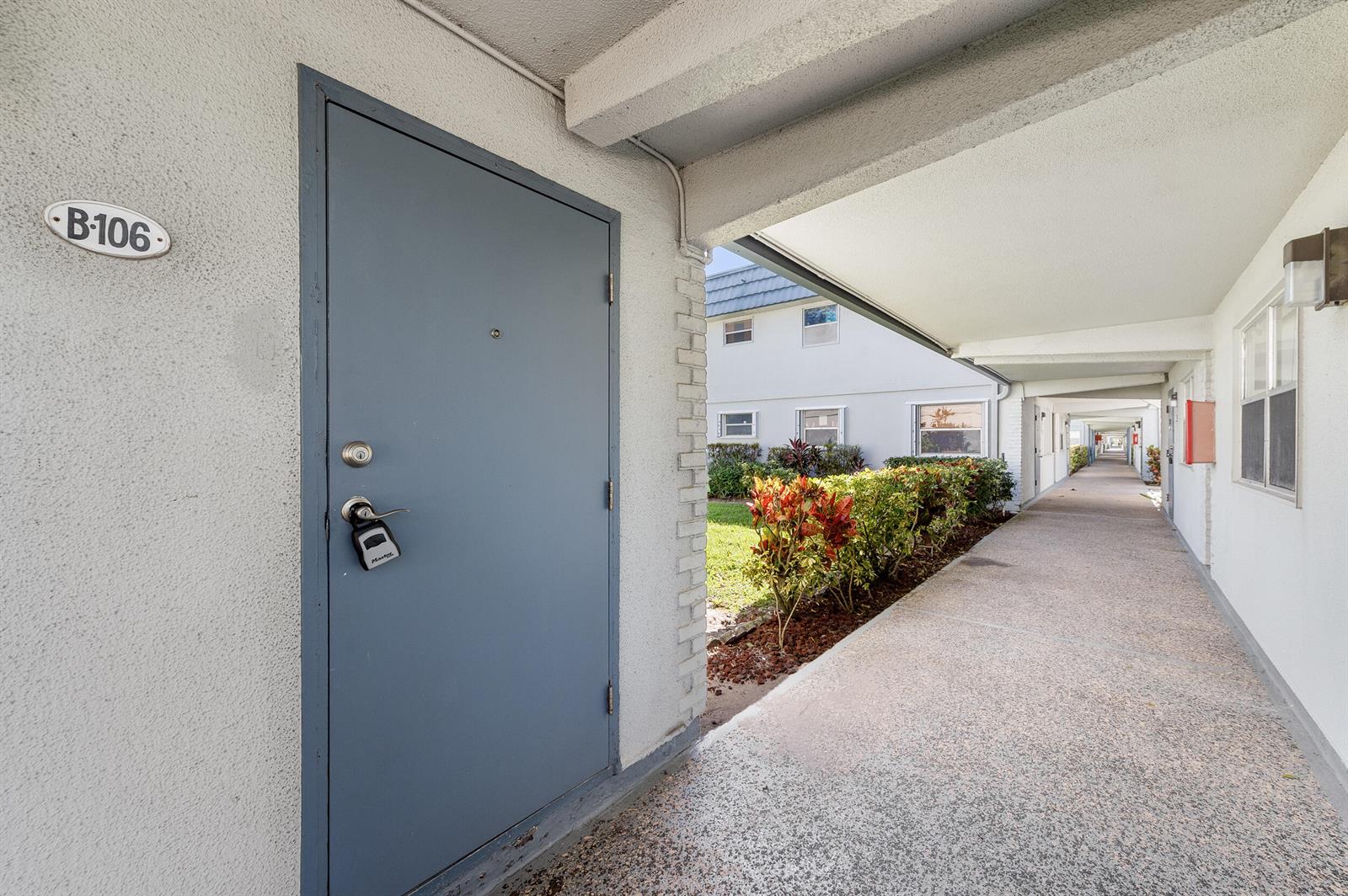 A gray door labeled B-106 opens onto a covered outdoor walkway beside a garden — 106 Tuscany B, Delray Beach FL 33446, DELRAY BEACH, FL