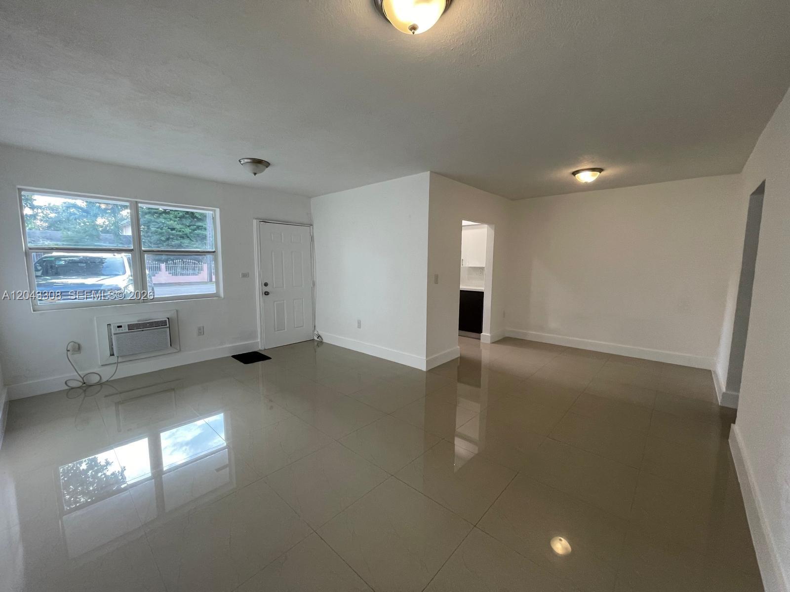 A bright, empty living room features white walls, large beige floor tiles, and a window unit — MIAMI, FL