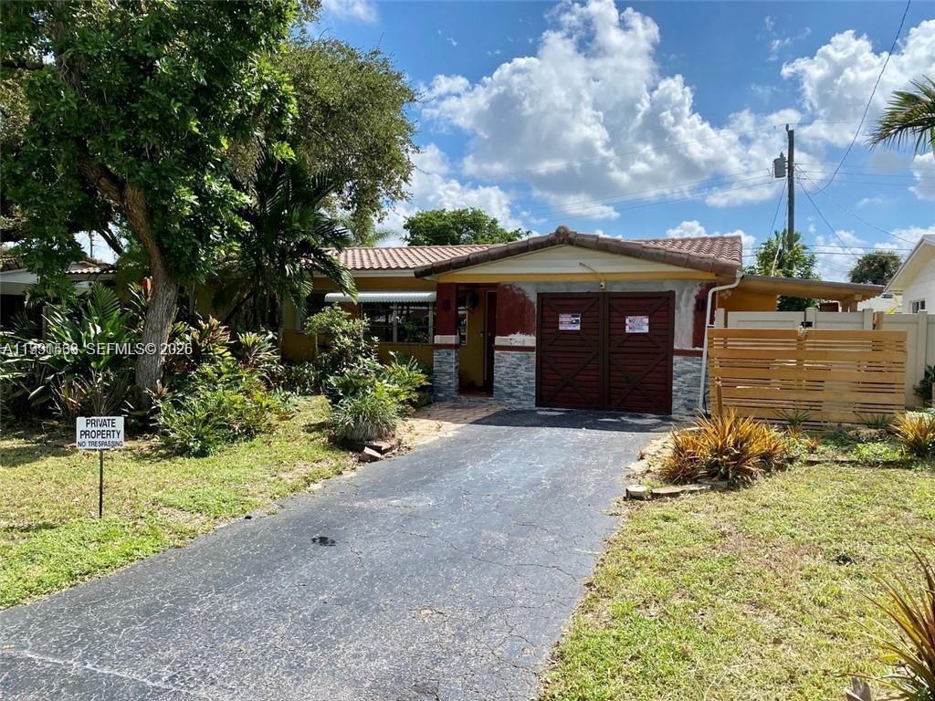 A single-story house with a paved driveway, a wooden garage door, and lush landscaping — 516 NE 28th Dr, Wilton Manors FL 33334, WILTON MANORS, FL