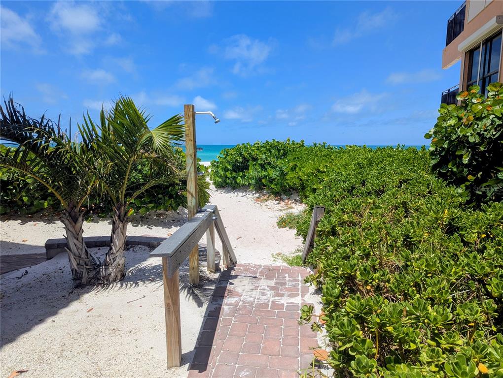 A brick walkway leads through lush greenery to a sandy beach path with an outdoor shower — 6006 GULF DRIVE UNIT 207, HOLMES BEACH, FL
