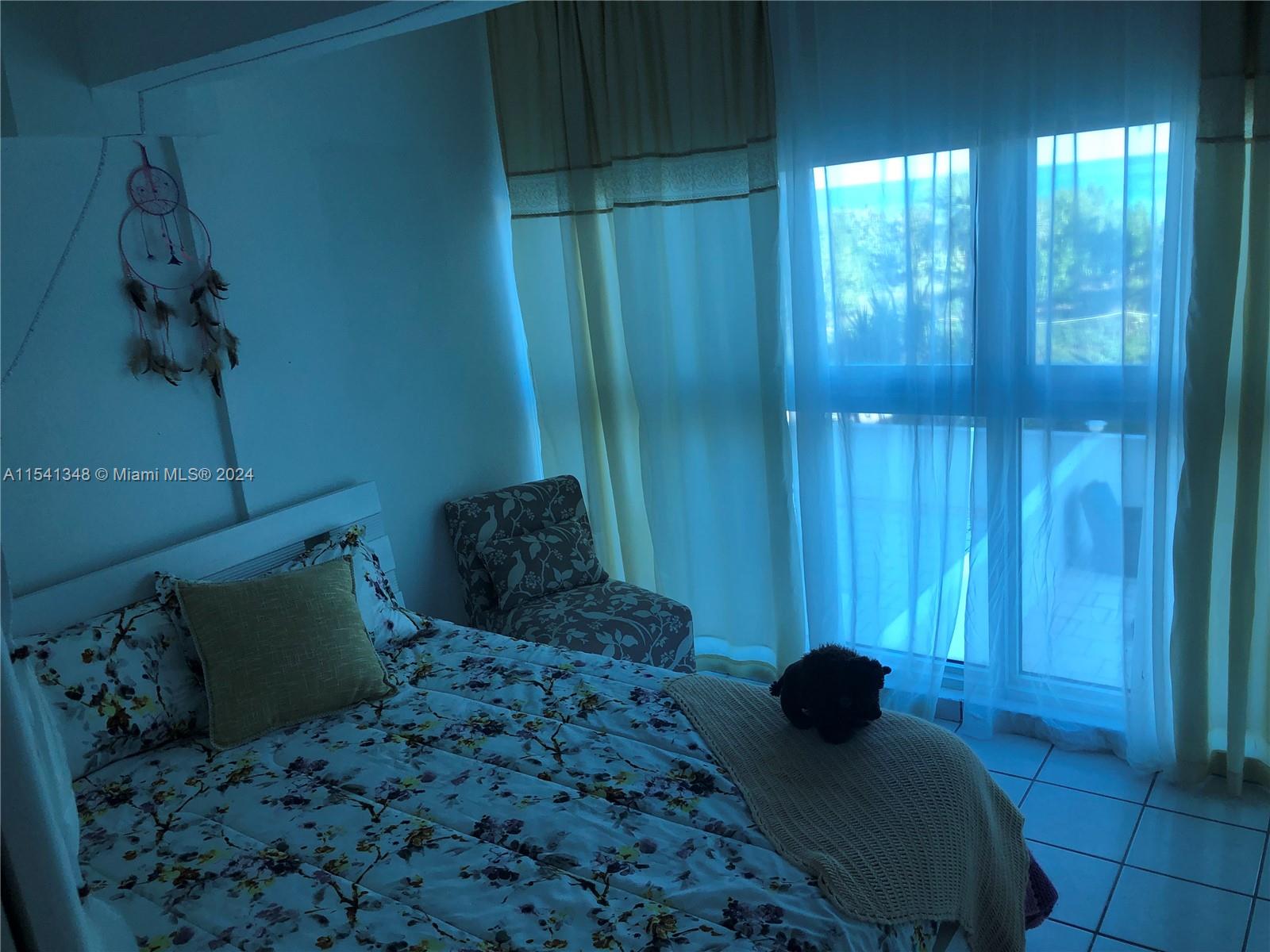 A bedroom features a floral bed, a patterned armchair, and sheer curtains covering a window — 5445 Collins Ave, Miami Beach FL 33140, MIAMI BEACH, FL