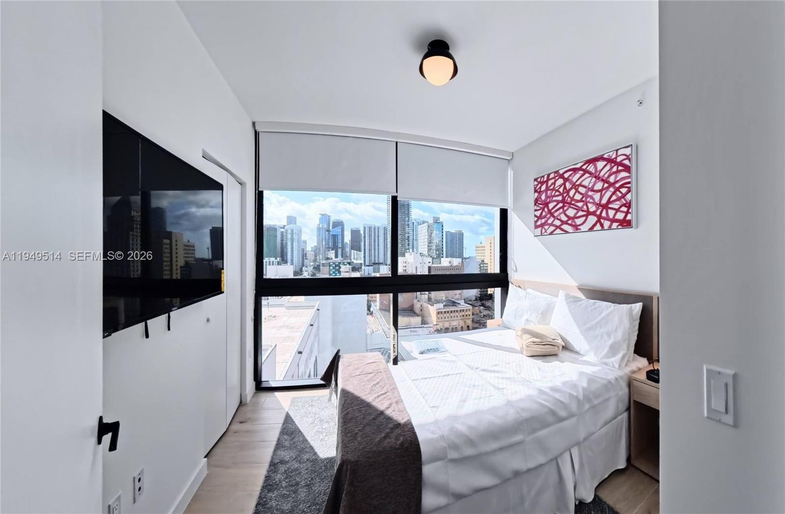 A modern bedroom features a bed, wall-mounted television, and floor-to-ceiling windows overlooking a city skyline — 225 N Miami Ave # 1812, Miami FL 33132, MIAMI, FL