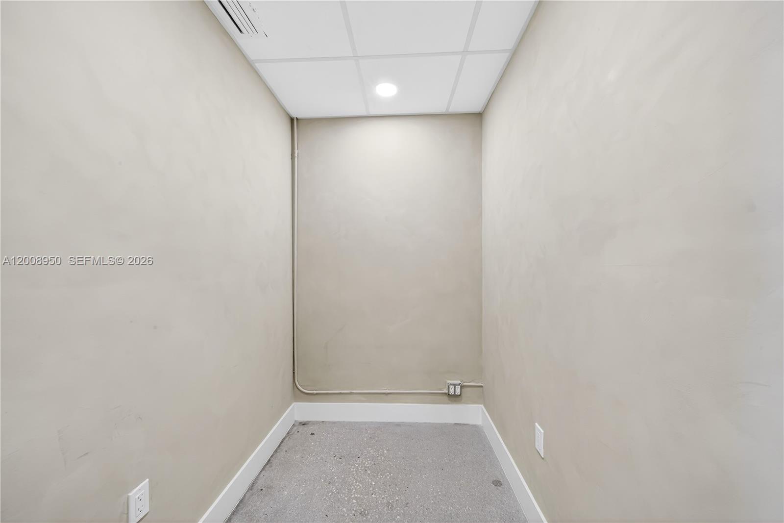 A small, empty room features beige walls, a drop ceiling with a recessed light, and concrete flooring — 12305 SW 133rd Ct # 26, Miami FL 33186, MIAMI, FL