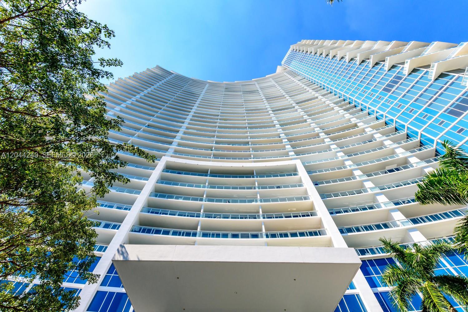 A low-angle view of a curved, modern high-rise building with glass balconies and blue windows — 2020 N Bayshore Dr # 3904, Miami FL 33137, MIAMI, FL