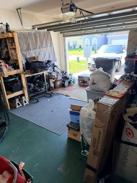 A cluttered garage features a workbench, storage shelving, a lawnmower, and a view of a driveway — 719 CORVINA DRIVE, DAVENPORT, FL
