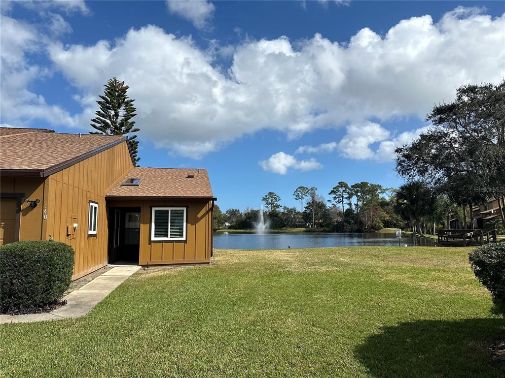 A brown exterior building sits on a grassy lawn beside a pond with a fountain — 156 LAKEWOOD VILLAGE CIRCLE, DAYTONA BEACH, FL