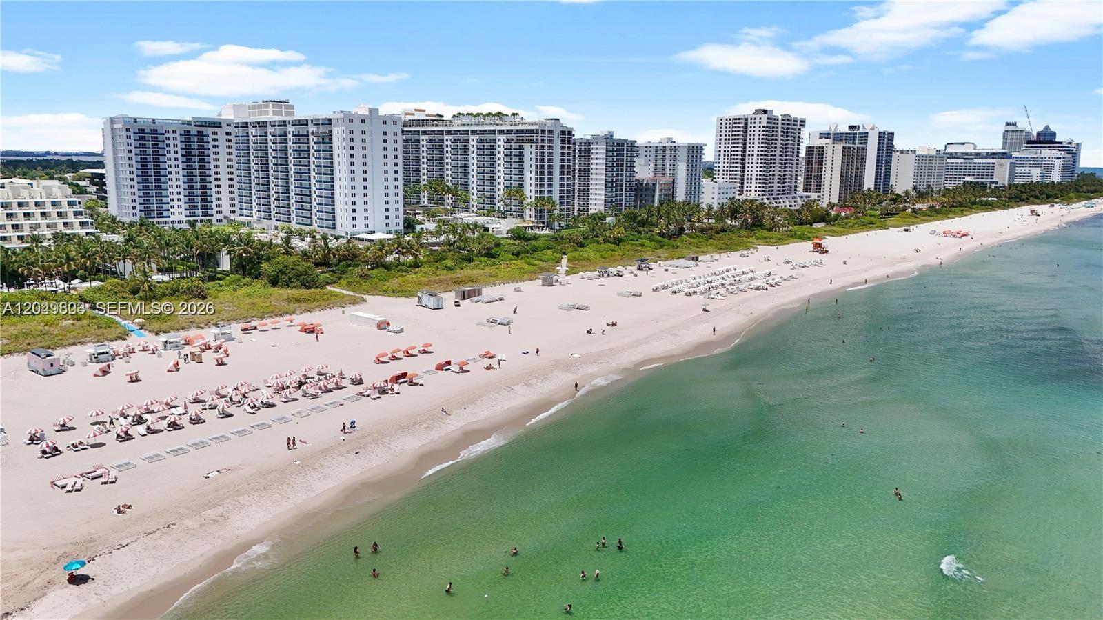 An aerial view of a sandy beach with numerous umbrellas, swimmers, and large residential buildings — 2301 Collins Ave # 414, Miami Beach FL 33139, MIAMI BEACH, FL