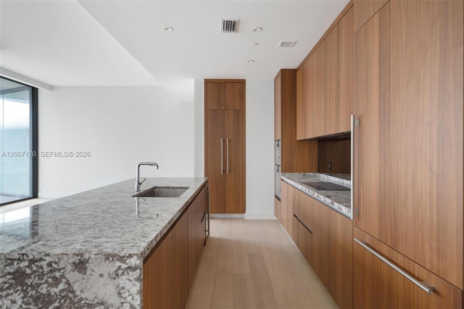 A modern kitchen features wood cabinetry, a granite-topped island with a sink, and light flooring — 175 SE 25th RD # 1904, Miami FL 33129, MIAMI, FL