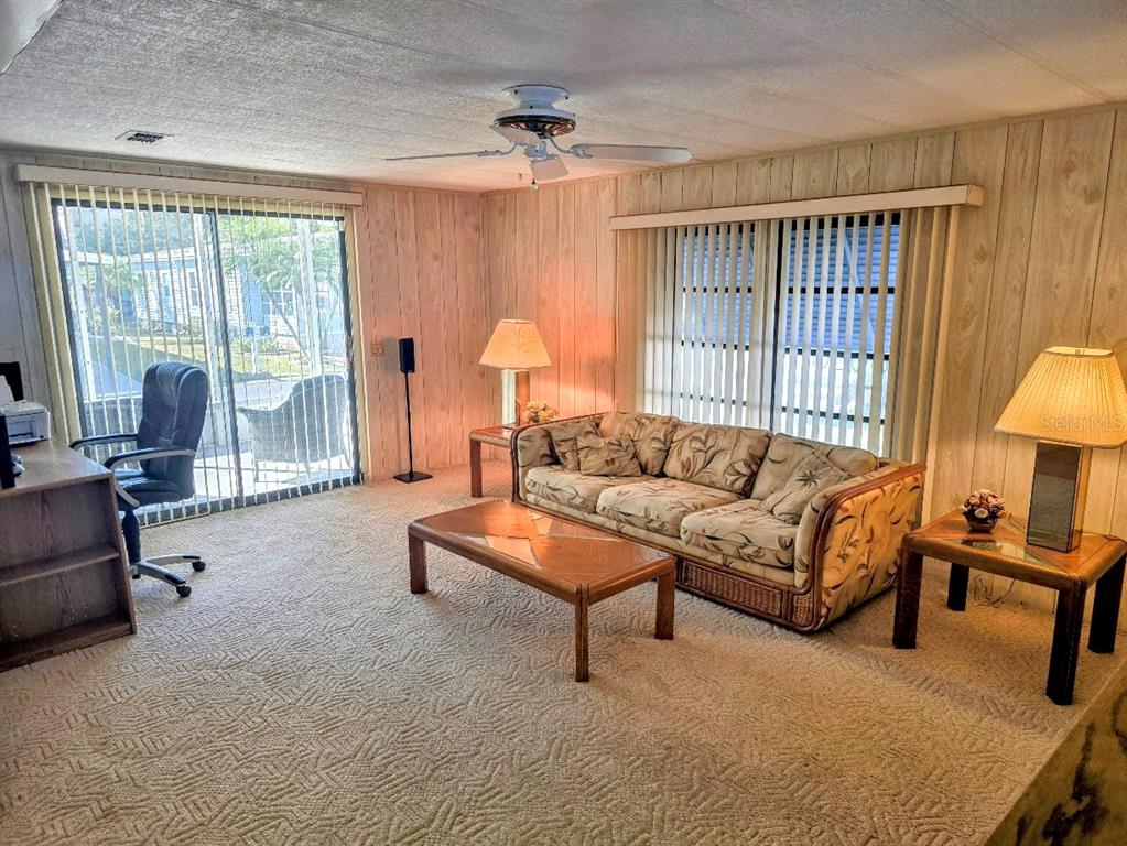 A living room with wood-paneled walls, beige patterned carpet, a sofa, and sliding glass doors — 12501 ULMERTON ROAD UNIT 188, LARGO, FL