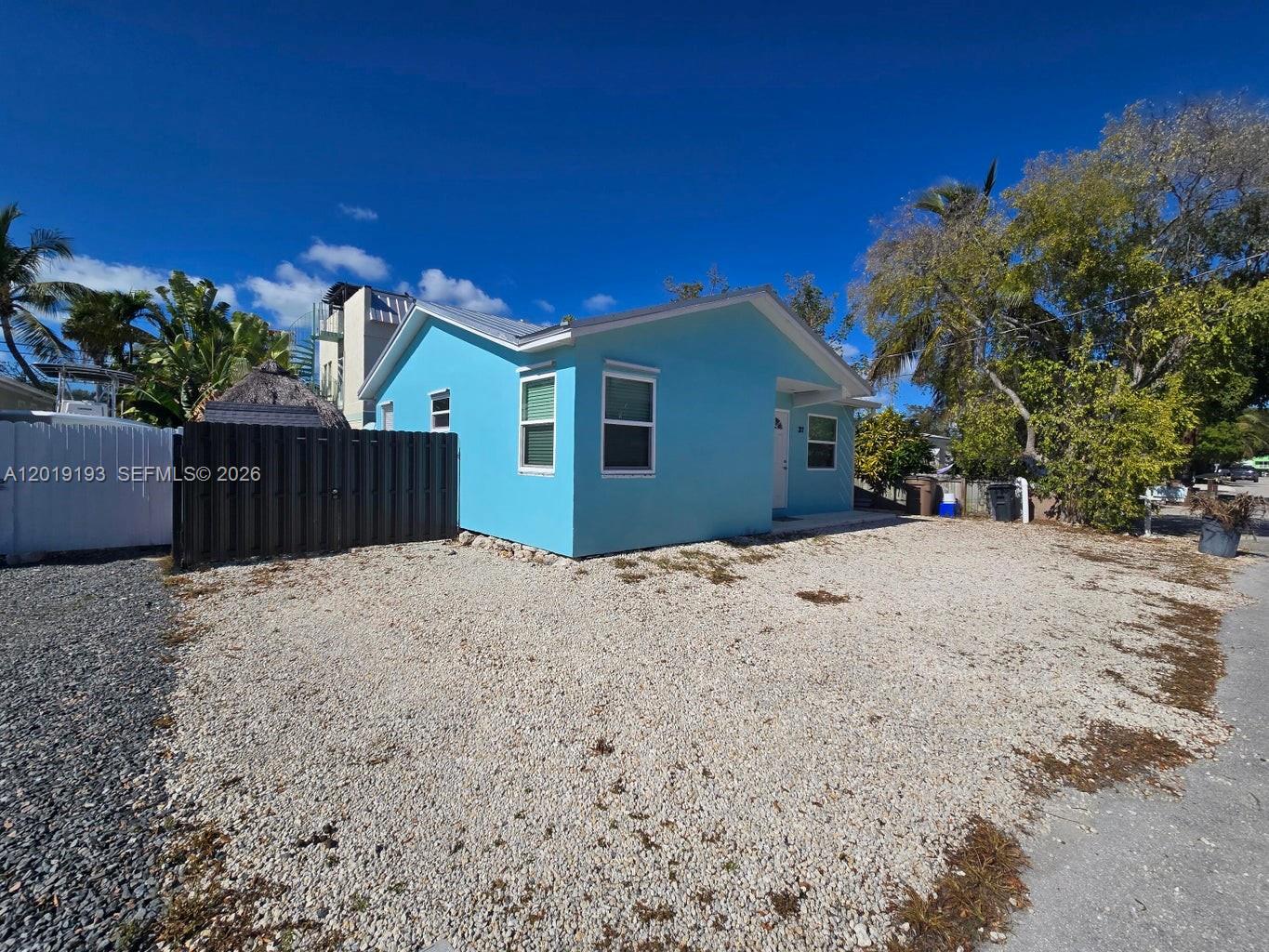 A light blue house with a gravel driveway and a dark fence under a blue sky — 37 Orange Dr, Key Largo FL 33037, KEY LARGO, FL